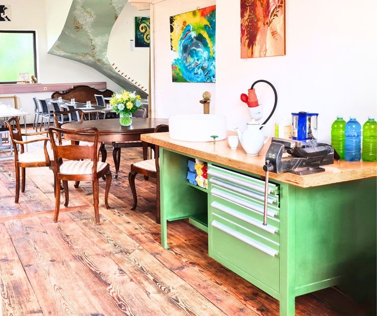 Innenraum einer Teeküche mit einem grünen Arbeitstisch, auf dem Teekannen, eine Kaffeemaschine und Wasserflaschen stehen, im Hintergrund ein runder Holztisch mit Stühlen und bunte Wandbilder an der weißen Wand.